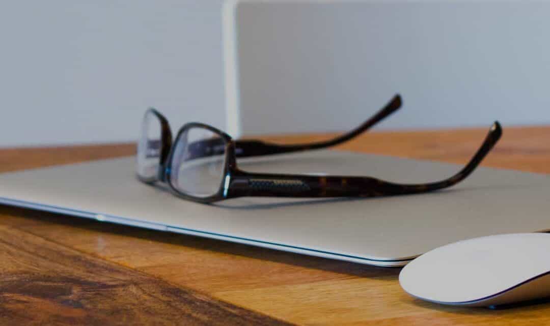 pair of spectacle glasses ontop of closed laptop with computer mouse on desk